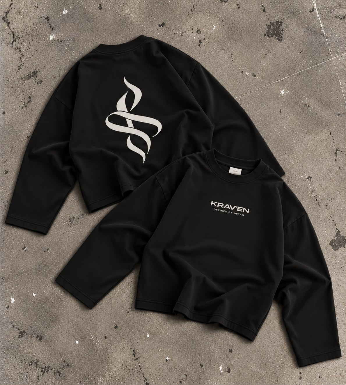 Krav'en Monogram Jumper in Black, front and back, laid flat on concrete: the ribbon monogram in bone across the back, the wordmark on the chest.